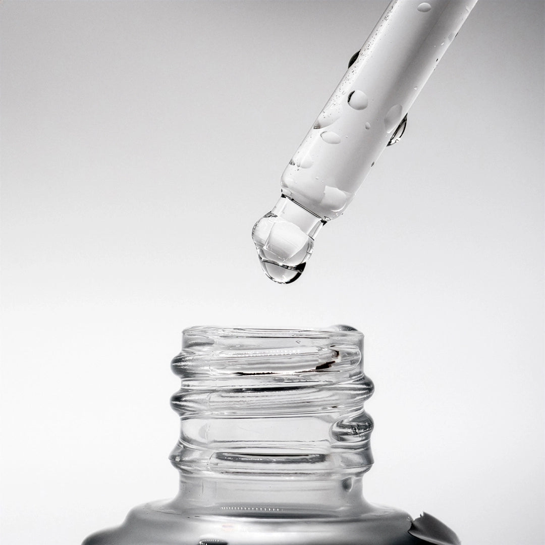 Glass dropper bottle with clear liquid being dispensed on a light gray background